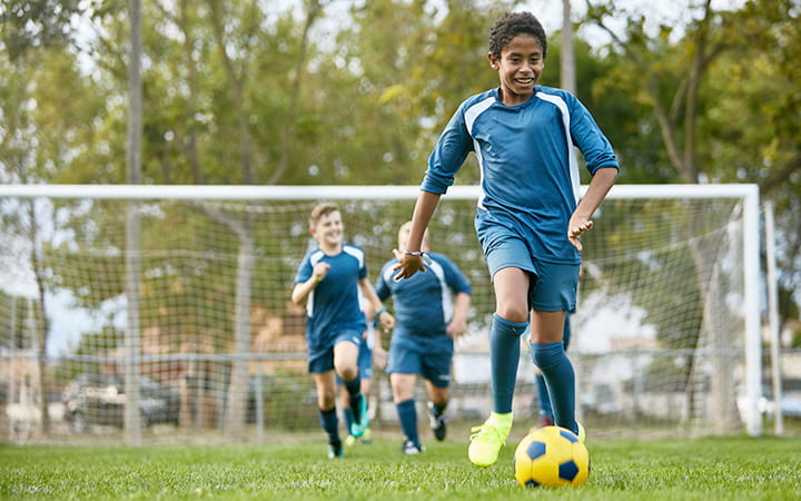 Smiling 13 year old male footballer dribbling ball toward camera with smiling teammates in pursuit in background