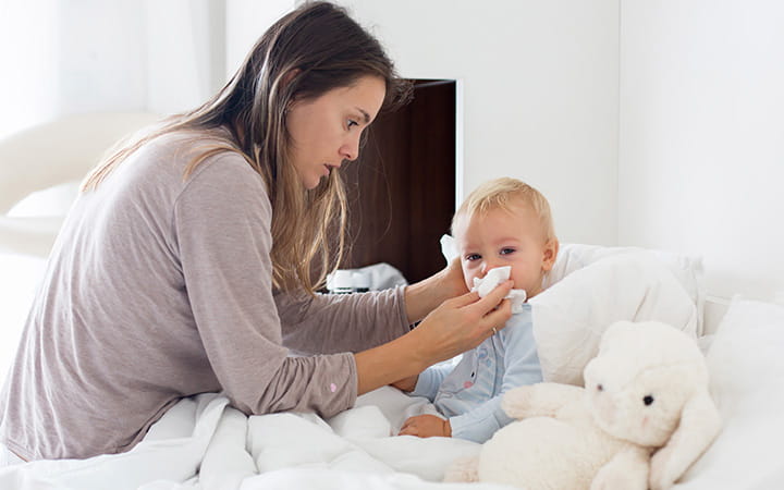 mother holds a tissue to a young toddler's nose in bed