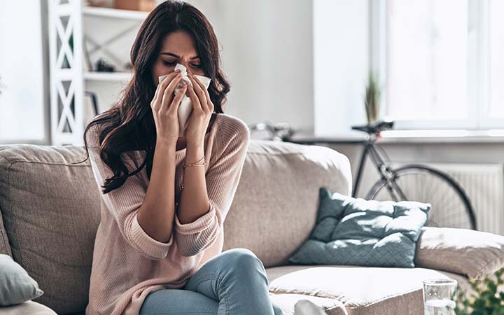 woman sitting on couch holding tissue to nose with both hands