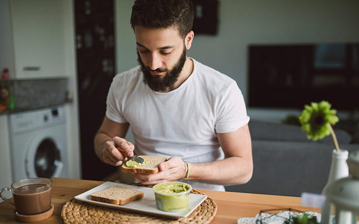 young man with beard spreading guacamole on bread seated in kitchen
