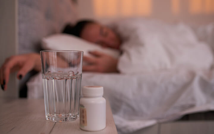 A jar with sleeping pills and a glass of water placed on bedside table near sleeping female