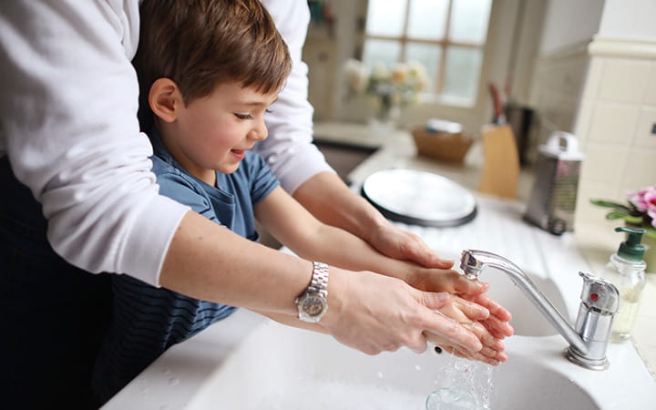 A four year old boy washing the dishes with his mum in the kitchen