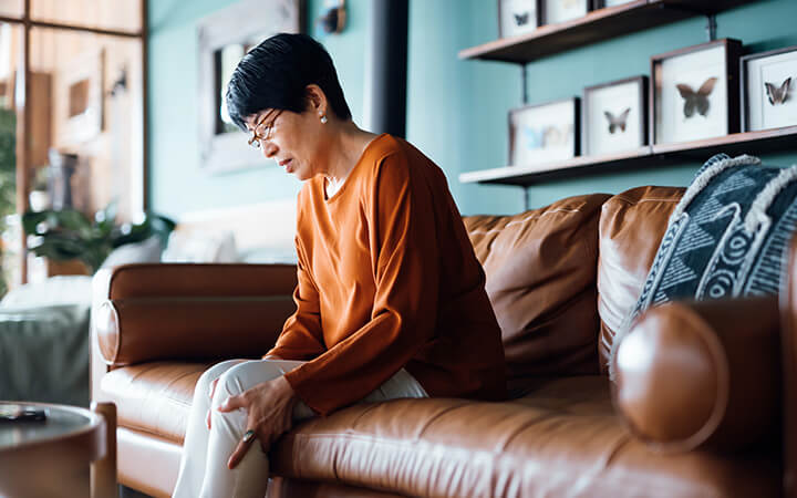 A distraught senior woman suffering from pain in leg while sitting on sofa in the living room at home