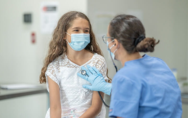 A girl sitting on the examination table at while her doctor uses a stethoscope to listen to her chest