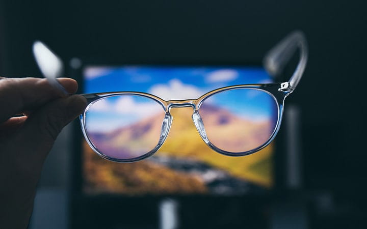 A pair of blue light computer glasses are held in front of a computer screen