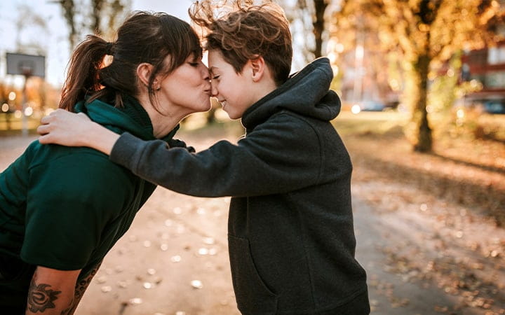 Young woman kissing and embracing her son outdoor, standing face to face on sunny day
