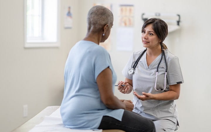 A middle-aged woman sits up on an exam table for a check-up with a nurse facing her
