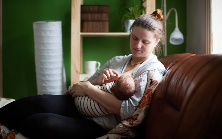 A mother feeds her newborn while sitting on a sofa at home