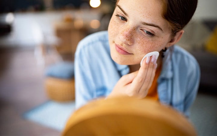 Teenage girl cleaning her face with cotton pad in front of the mirror