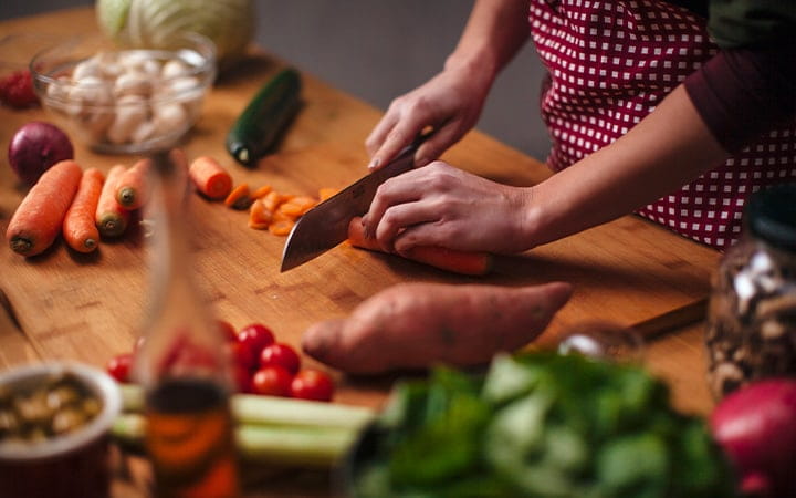 Hands of a young woman as she chops carrots with a big knife with various vegetables on the kitchen counter
