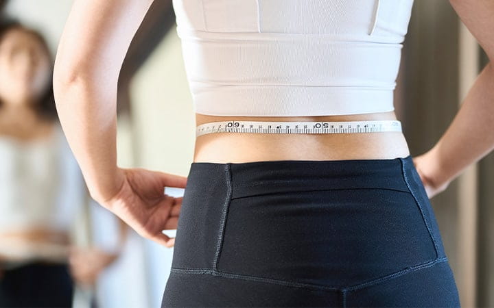 A woman holds a tape measure around her waist, checking diet results in a full-length mirror