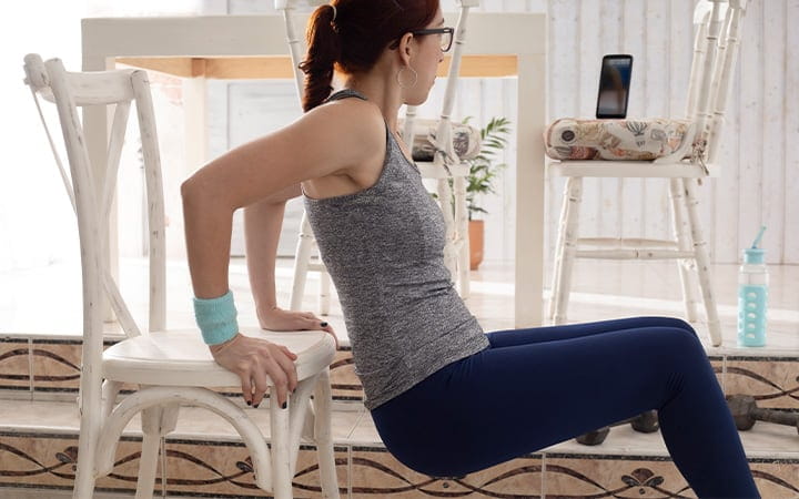 A woman practicing triceps on the dining room bench in a video conference from her cell phone