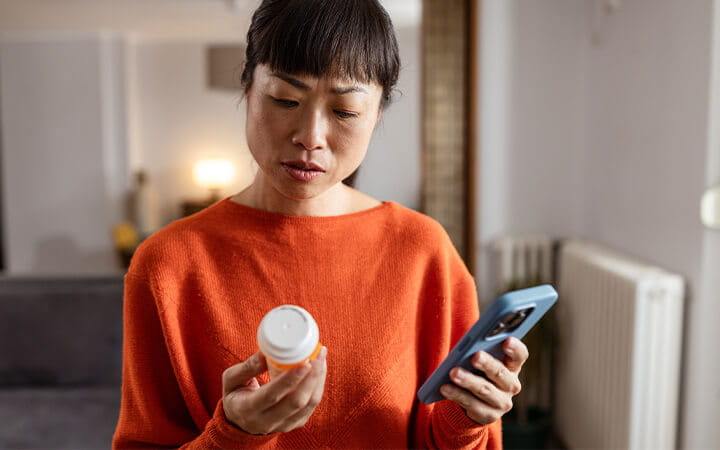 A serious mature woman is reading a pill bottle label and using a mobile phone at her home