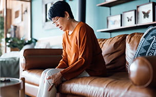 A distraught senior Asian woman feeling unwell, suffering from pain in leg while sitting on sofa in the living room at home