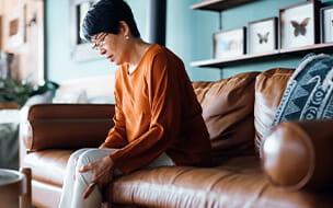 A distraught senior Asian woman suffering from pain in leg while sitting on sofa