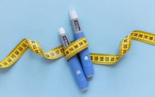 Two injectors dosing pens for subcutaneous injection wrapped in yellow measuring tape over a blue background