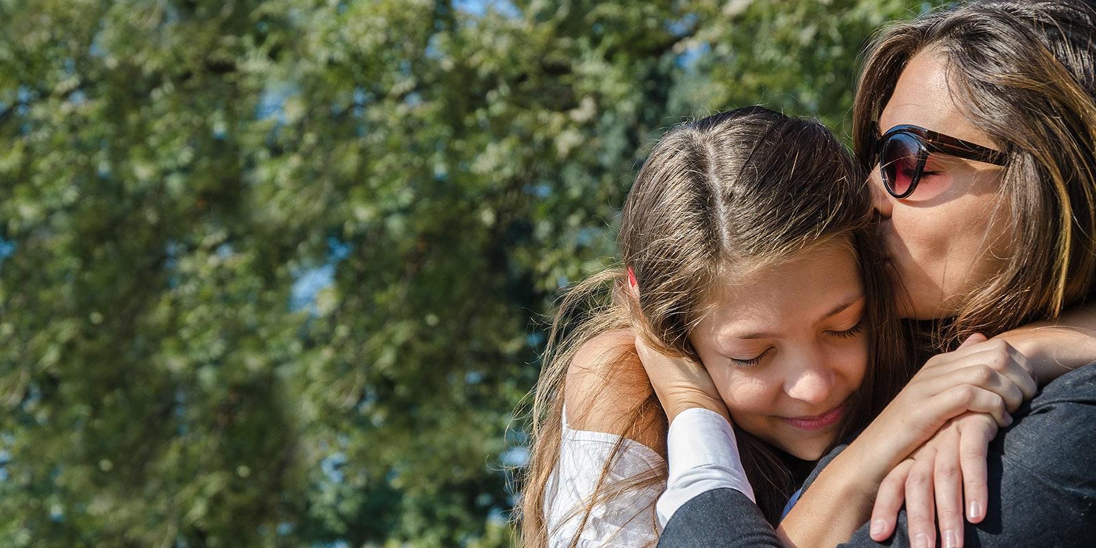 Mother kissing teen daughter's head smiling outside.