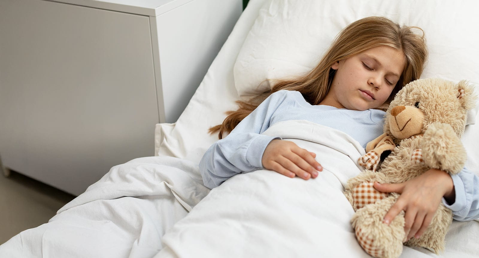 A child is peacefully resting in a hospital bed with a soft teddy bear for comfort and support