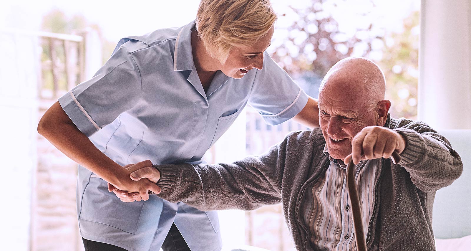 A female healthcare worker is supporting an elderly man, who is using a cane to stand up from his chair