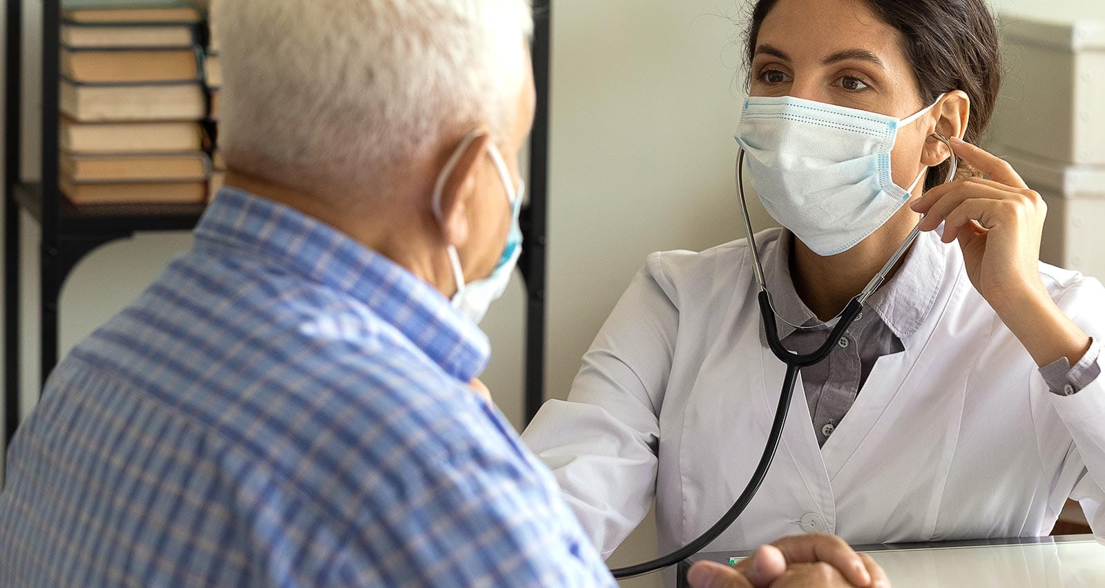 A female doctor in acfacemask listens to a male heart patient's chest with a stethoscope