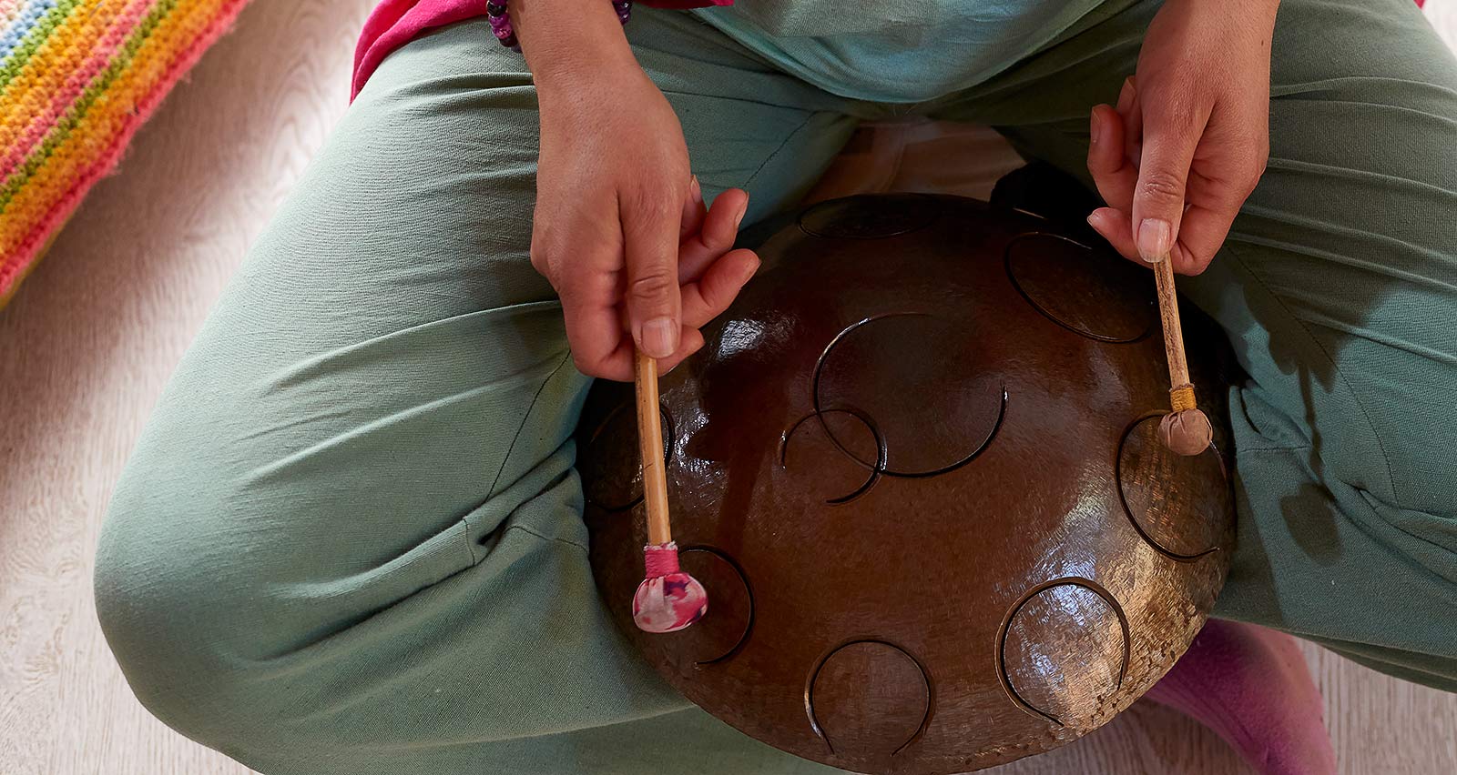 A woman is seated on floor, playing a steel tongue drum, with a Tibetan sound bowl nearby