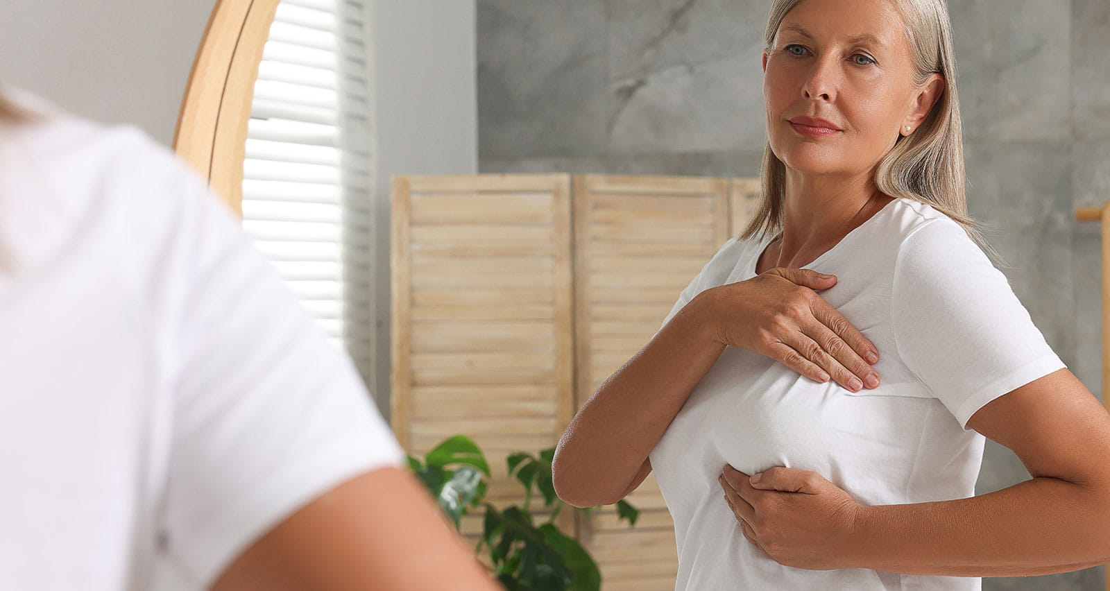 A senior woman performing a breast self-examination near a bathroom mirror