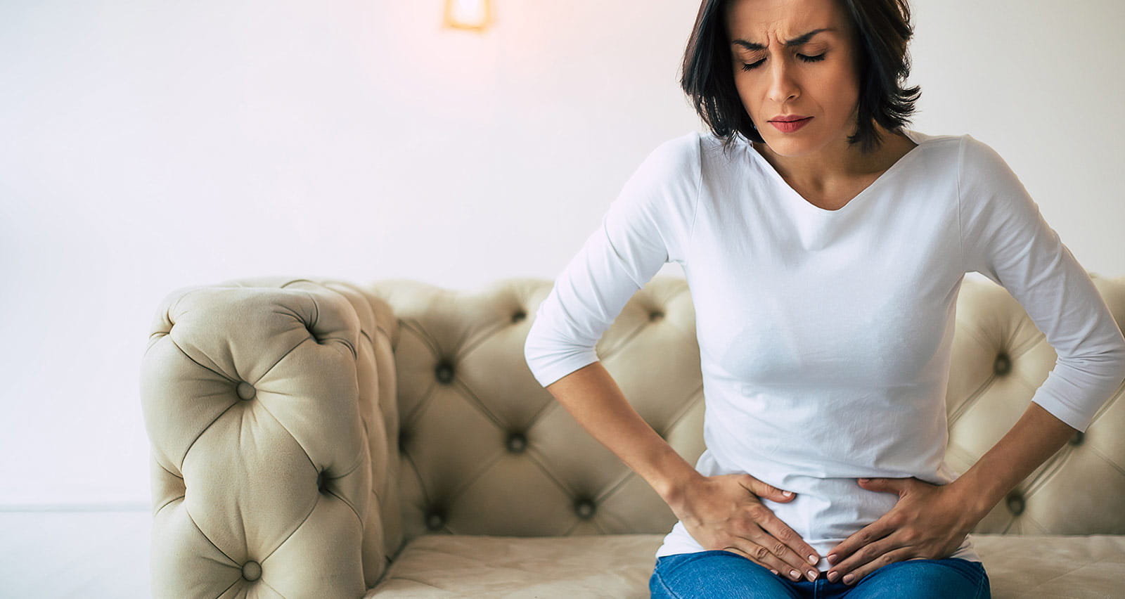 An adult woman is sitting on a sofa at home and holding her lower stomach while suffering from cramps