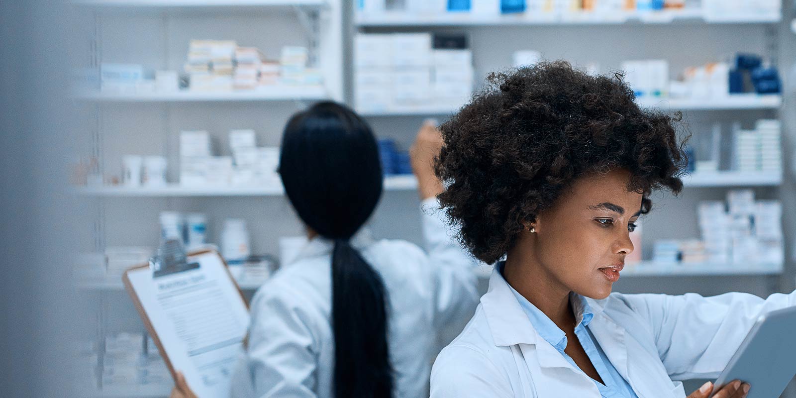 A young pharmacist doing inventory in a pharmacy on a digital tablet with her colleague in the background
