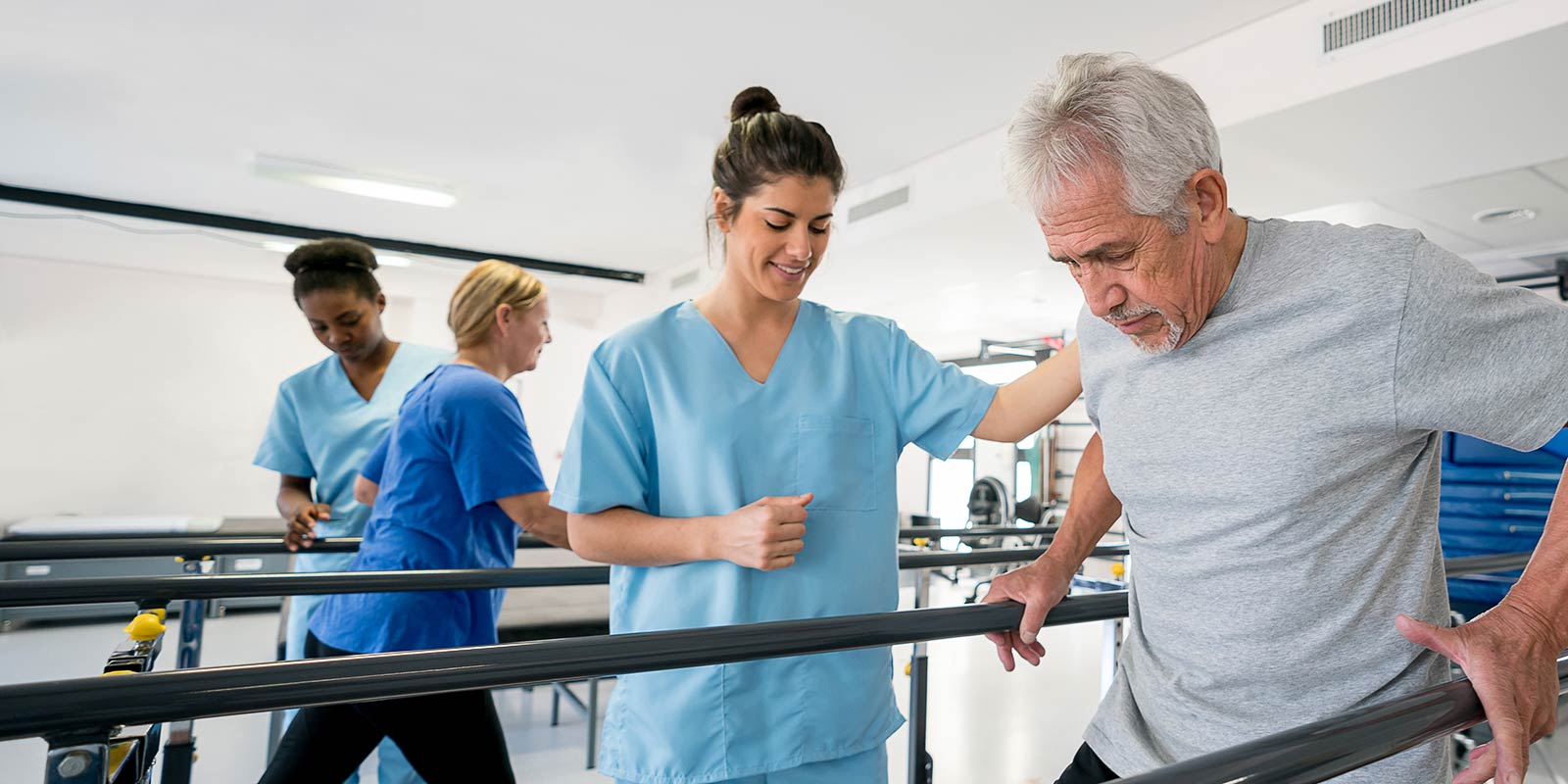 Diverse team of physiotherapist helping senior patients walk between parallel bars all looking happy and smiling